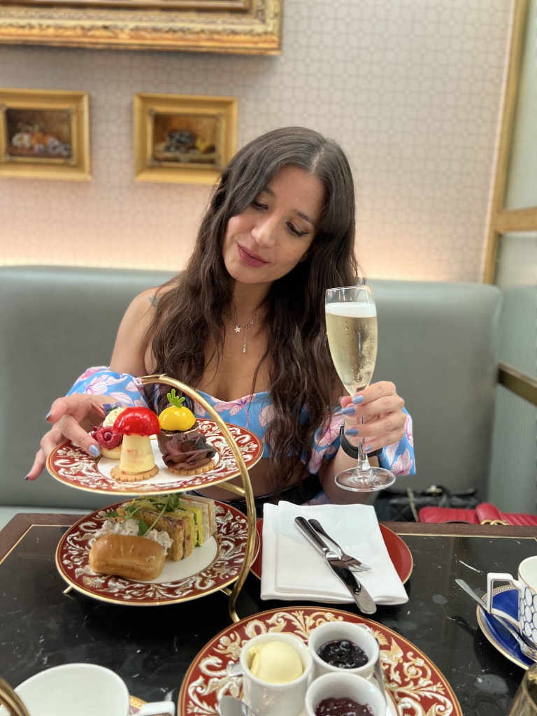 Dani holds a glass of champagne and stares lovingly at a two tier cake stand with sandwiches and pastries.