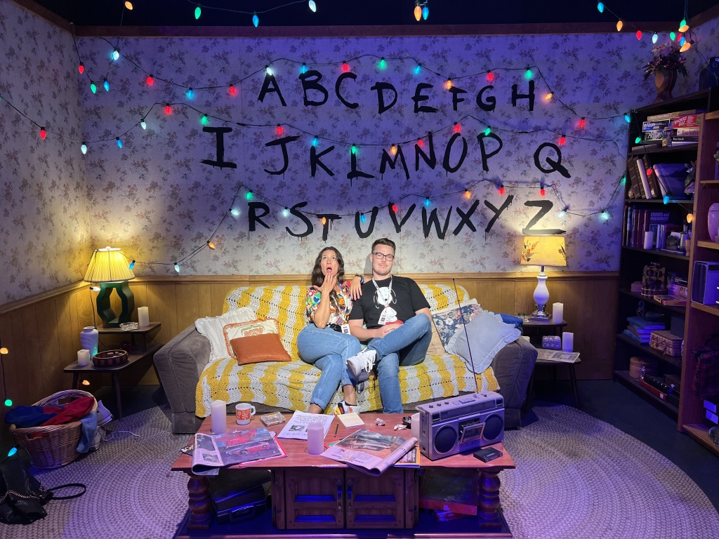 Dani and Dan sit on Joyce's sofa, with the alphabet and lights behind them at the #StrangerThingsExperience