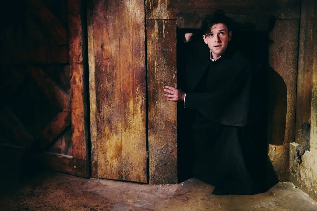A male actor wearing a black cape crouches by a small door on the floor. 